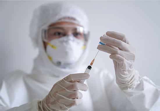 A medical worker in protective clothing fills a syringe with a substance from a bottle