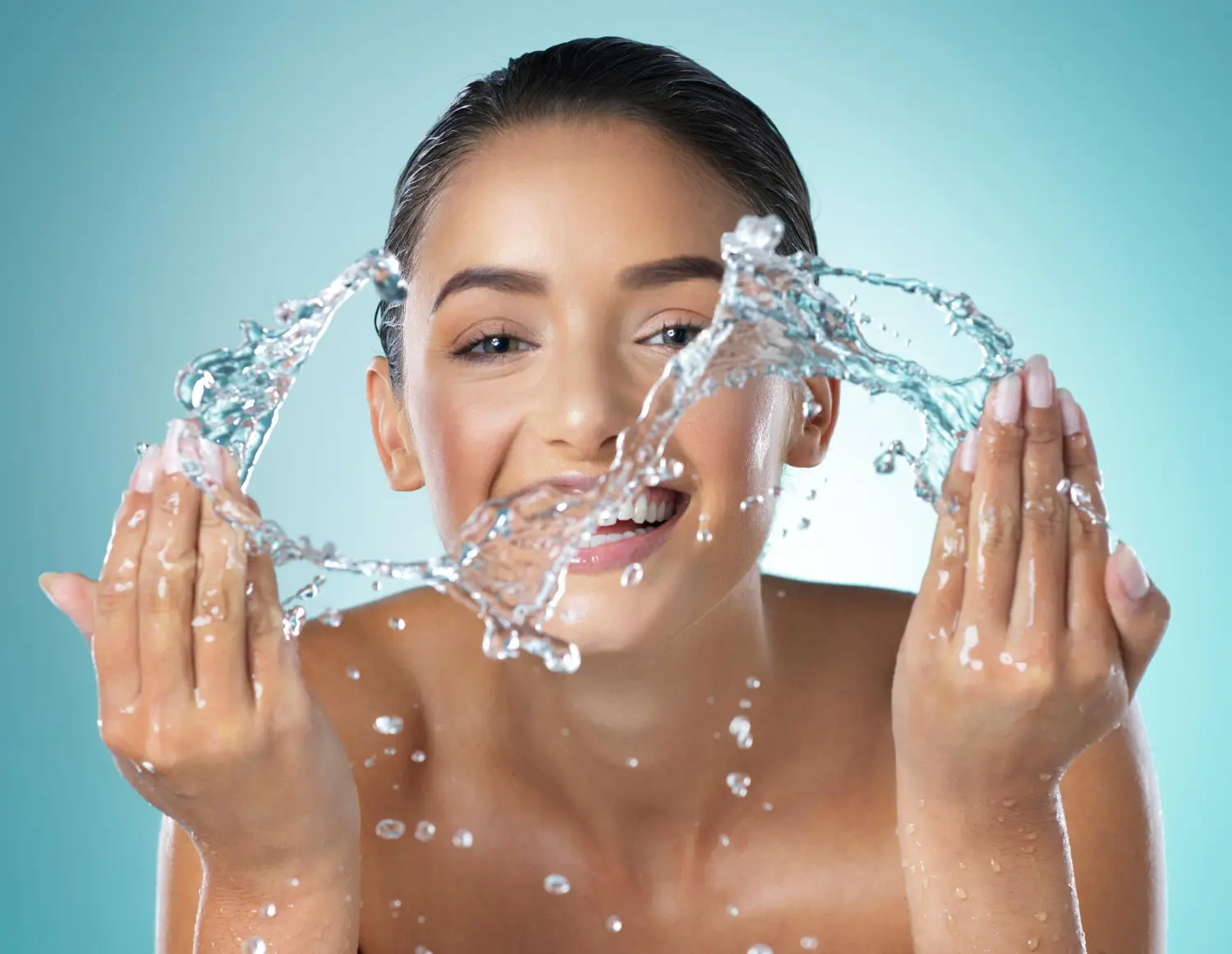 Beautiful young woman smiling while splashing water on her face, highlighting fresh skin and hydration.