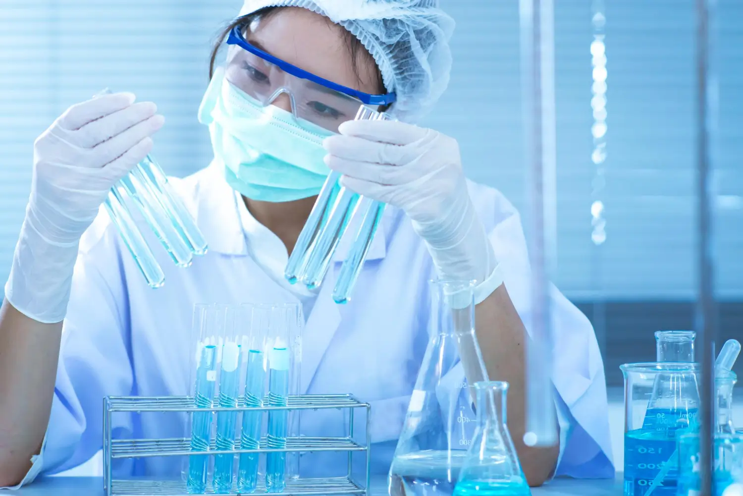 A focused female scientist in a lab coat, mask, and goggles examining test tubes with a blue liquid.