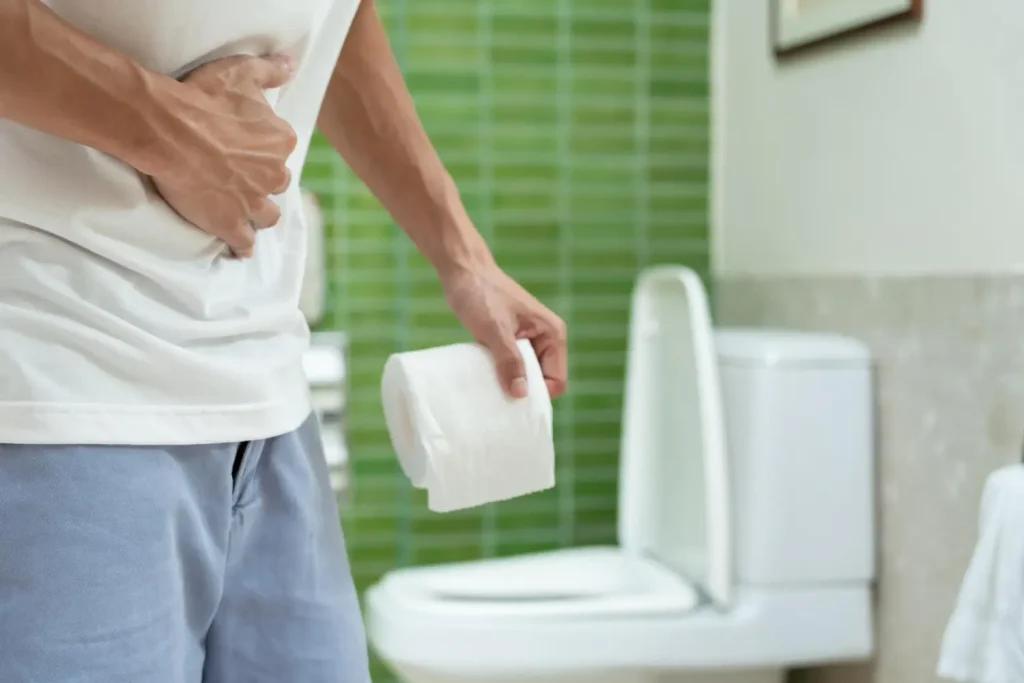 Man experiencing stomach pain holding toilet paper while standing in a bathroom, possibly experiencing a side effect of Mounjaro.
