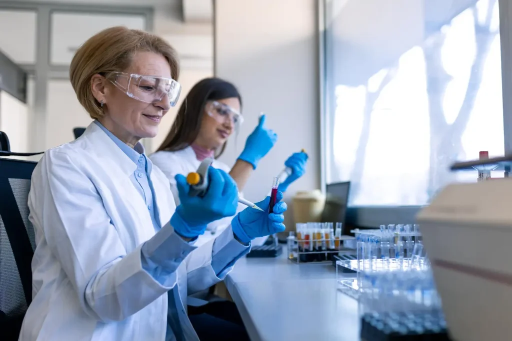 Two scientists in lab coats using pipettes and test tubes in a bright, modern laboratory setting.
